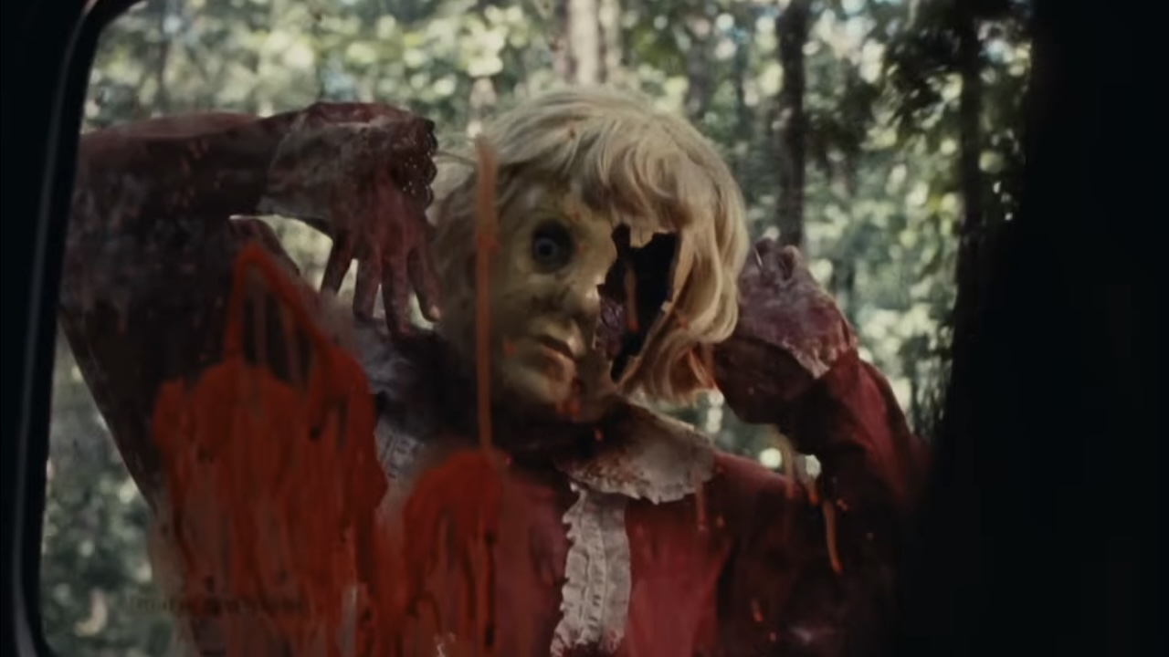 Doll-masked killer with broken eye section looking through bloody car window in Dolly