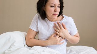 woman sitting up in bed with hands over heart, as if in discomfort