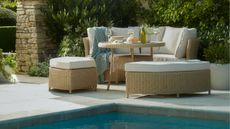 Outdoor Sorrento acacia wood dining table from Cox & Cox with curved seats and cushions, with additional backless seats by a pool on a tiled patio area. There is sand colored brickwork and evergreen foliage behind