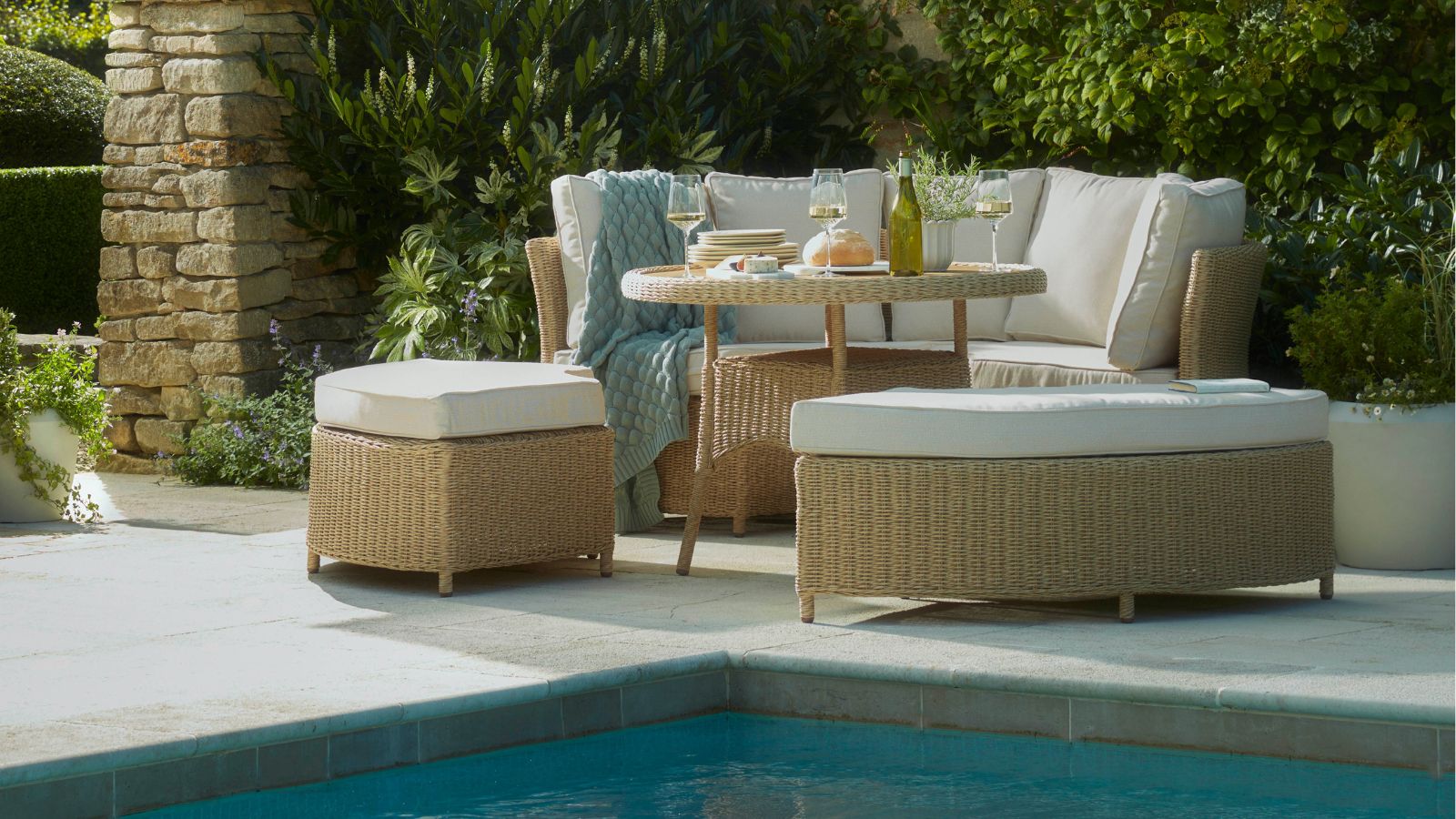 Outdoor Sorrento acacia wood dining table from Cox &amp; Cox with curved seats and cushions, with additional backless seats by a pool on a tiled patio area. There is sand colored brickwork and evergreen foliage behind