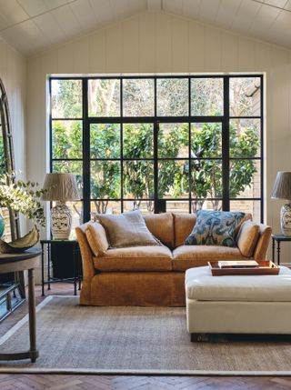 cream living room with shiplap walls, ochre brown couch, wooden floor, rug, footstool, side tables, crittall doors to outside