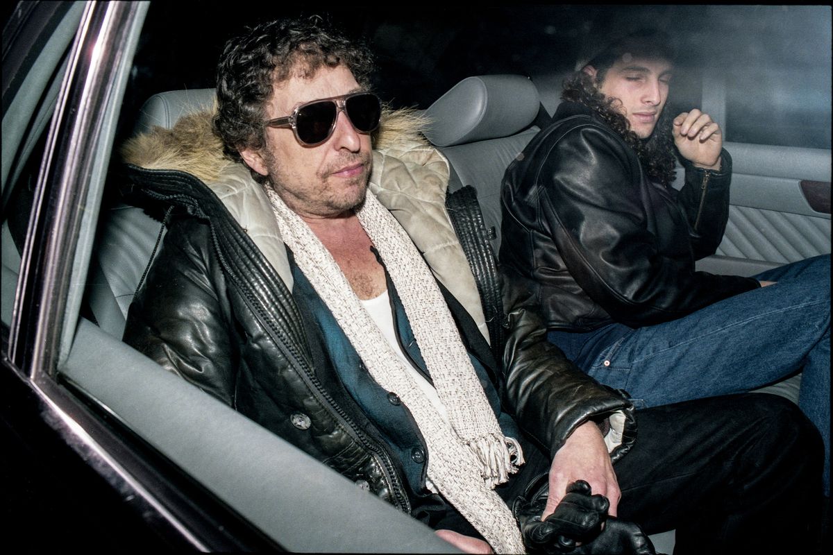 Colour photo of Bob Dylan in sunglasses, a fur-trimmed leather coat and white scarf in the back of a car, with his son Jakob Dylan turning away beside him.