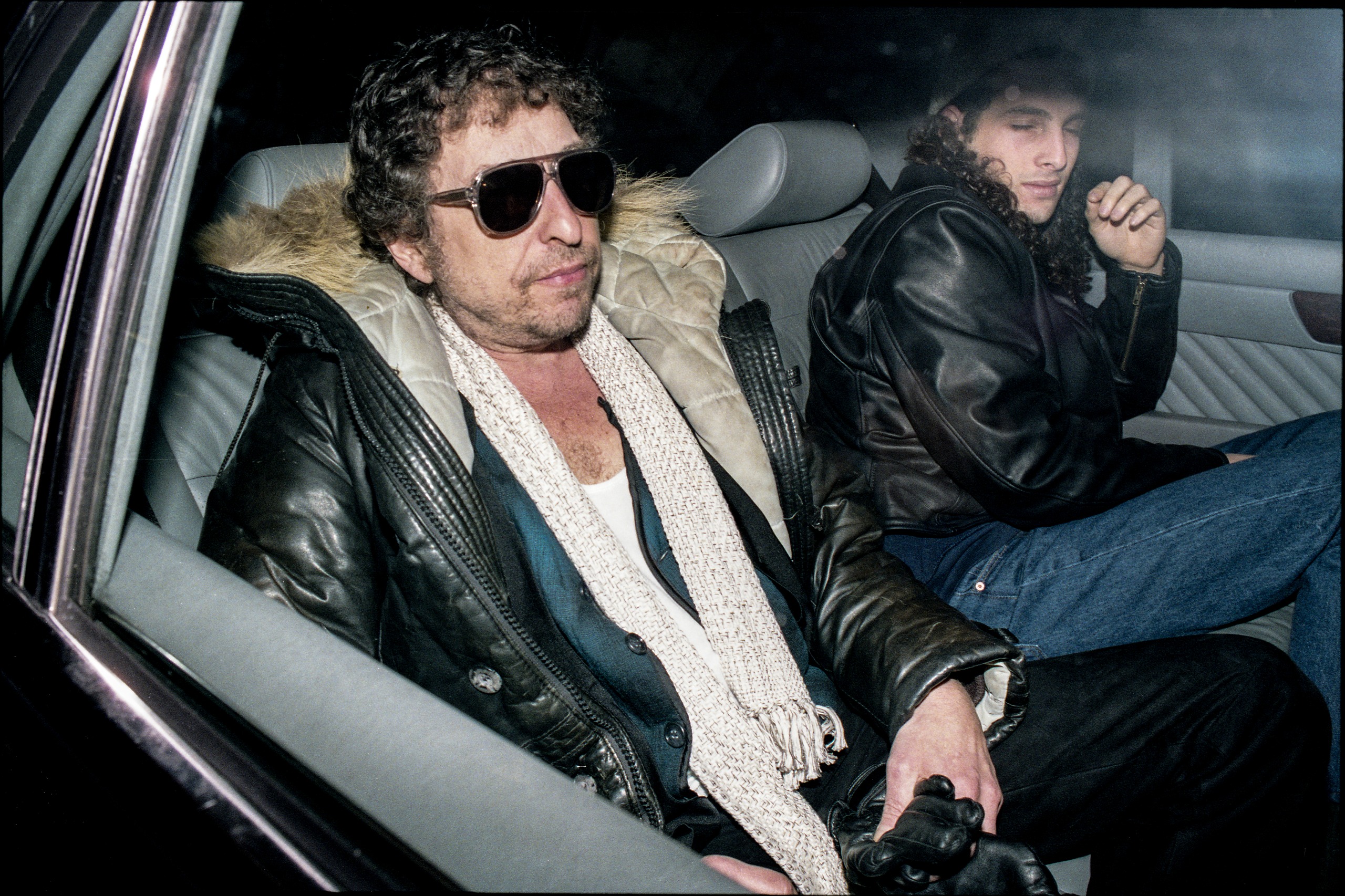 Colour photo of Bob Dylan in sunglasses, a fur-trimmed leather coat and white scarf in the back of a car, with his son Jakob Dylan turning away beside him.