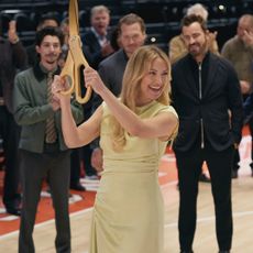 A woman (Kate Hudson as Isla Gordon) holds up giant gold scissors as a crowd (l-r Fabrizio Guido as Jackie, Scott MacArthur as Ness Gordon, Justin Theroux as Cam Gordon) stand behind her on a basketball court, in 'Running Point' season 2.