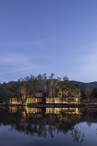The exterior of the ranch photographer at night. The ranch sits at the edge of a lake, surrounded by trees and mountains in the back. The walls are covered in glass and we see the lights on.