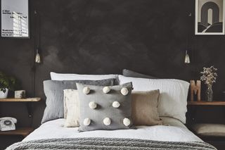 Bed with white bed covers and pom pom cushion against a dark concrete-effect wall, with handmade wooden shelves as bedside storage