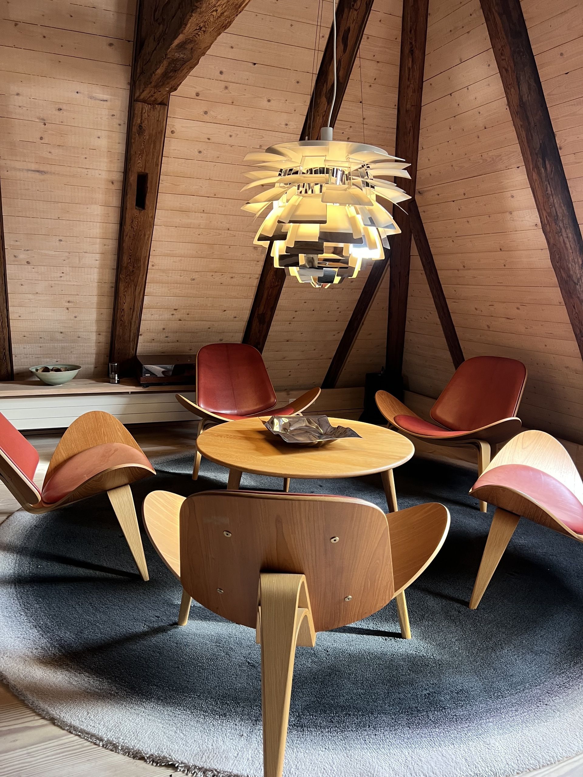 Image of five red leather and wood shell chairs around a coffee table on top of a blue rug, with a metal pendant light overhead.