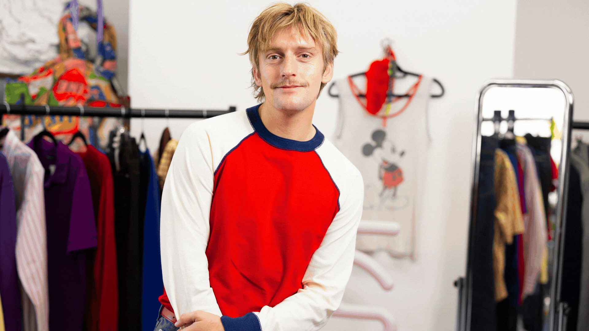 A rotating gif showing a young blonde man in a red, blue, and white long-sleeved jumper, standing in a filled-with-colorful-clothing studio, interspersed with images of whimsical designs.