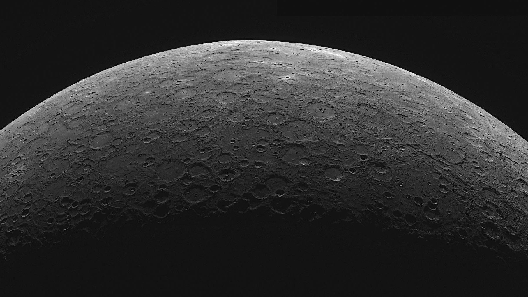 An image showing the upper curve of a barren grey planet against the blackness of space. The lower section of the image is shadowered and many craters can be seen pockmarking the surface.