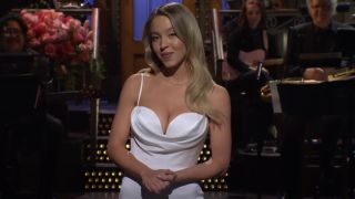 Sydney Sweeney holding her hands together in front of her and almost smiling during her SNL monologue.