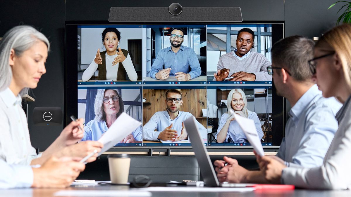 A corporate meeting taking place in front of a display with many happy faces in a videoconference meeting.