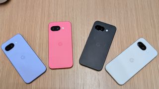 The Iris, Peony, Obsidian, and Porcelain Google Pixel 9a arrayed in a rainbow on a brown desk surface.