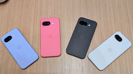 The Iris, Peony, Obsidian, and Porcelain Google Pixel 9a arrayed in a rainbow on a brown desk surface.