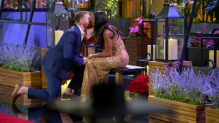 Stephen Richardson, on bended knee, and Monica Davis, sitting on a bench, kiss in a room filled with lavender planters, candles, and roses, in 'Love is Blind' season 7.