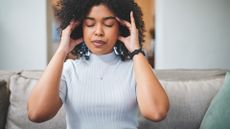 woman sat on a sofa with fingers in her ears and on her temples with eyes closed. 