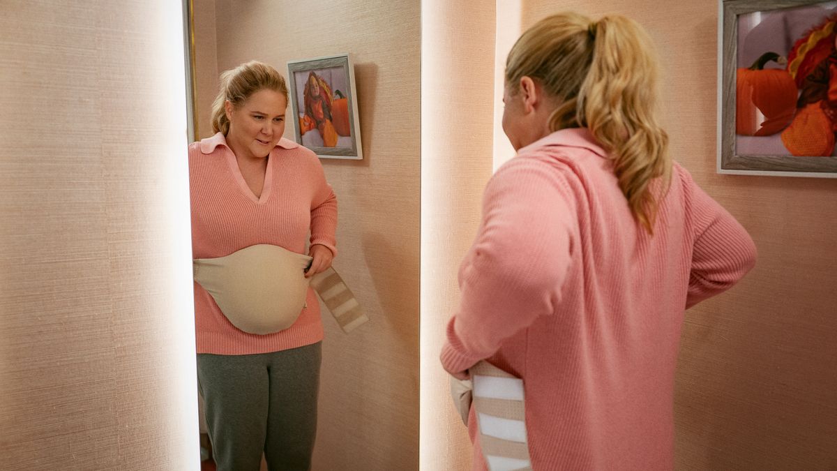 Lainy straps on a fake baby bump in front of a mirror in Kinda Pregnant.