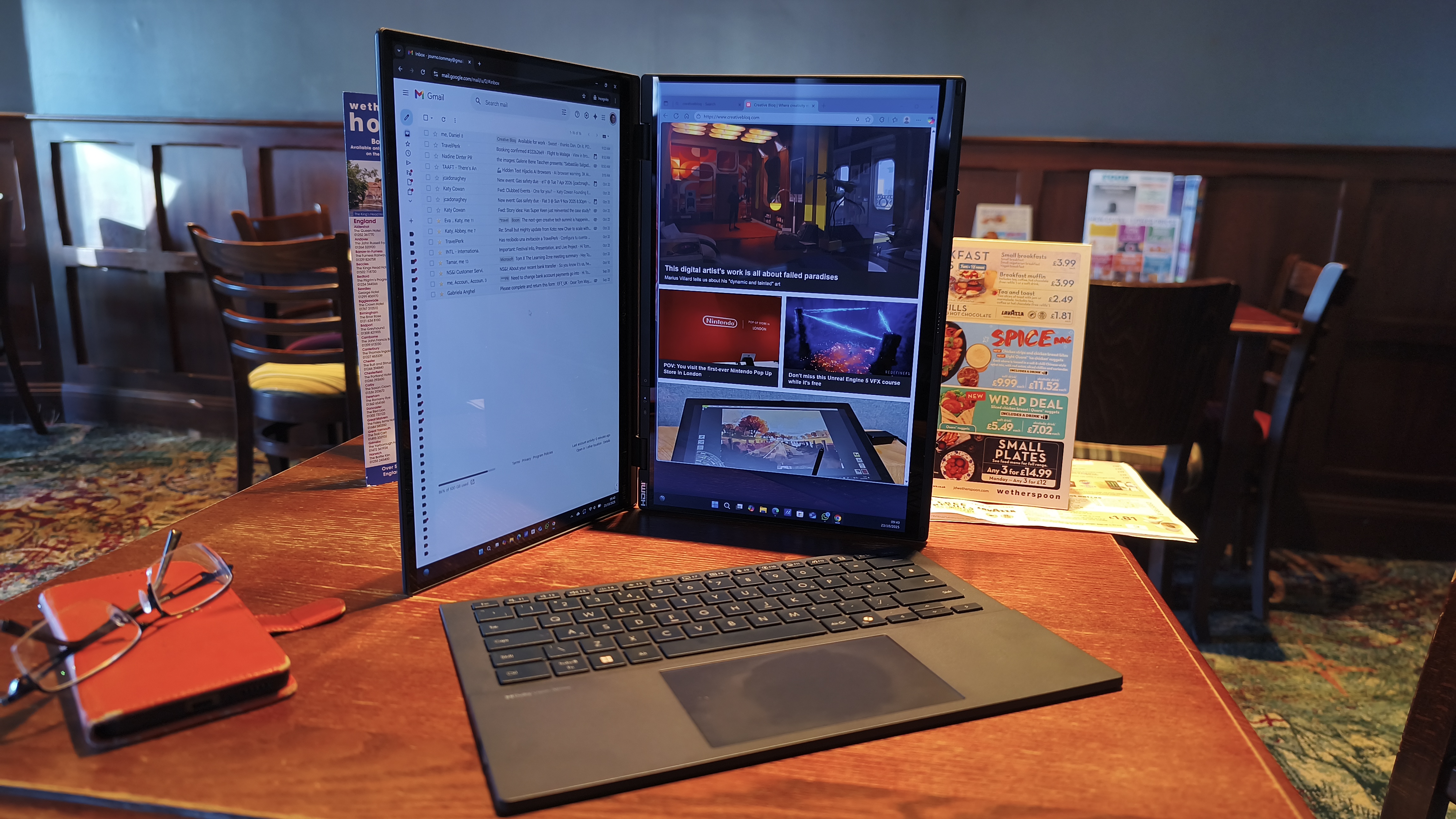 A dual-screen laptop is set up in a vertical book-like orientation with a detached keyboard and trackpad resting in front of it on a pub table.