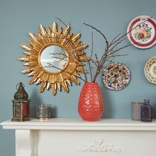 A white fireplace mantel set against a blue wall with a vase of seasonal branches and decorative plates hung on the wall