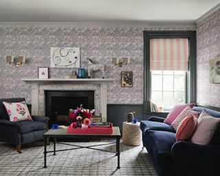 Living room with dado atelier wallpaper, blue painted paneling and wooden features, blue sofa and armchair, pink and red cushions, black metal coffee table, marble fireplace, pink an white striped blind, two shade wall lamps and wall art