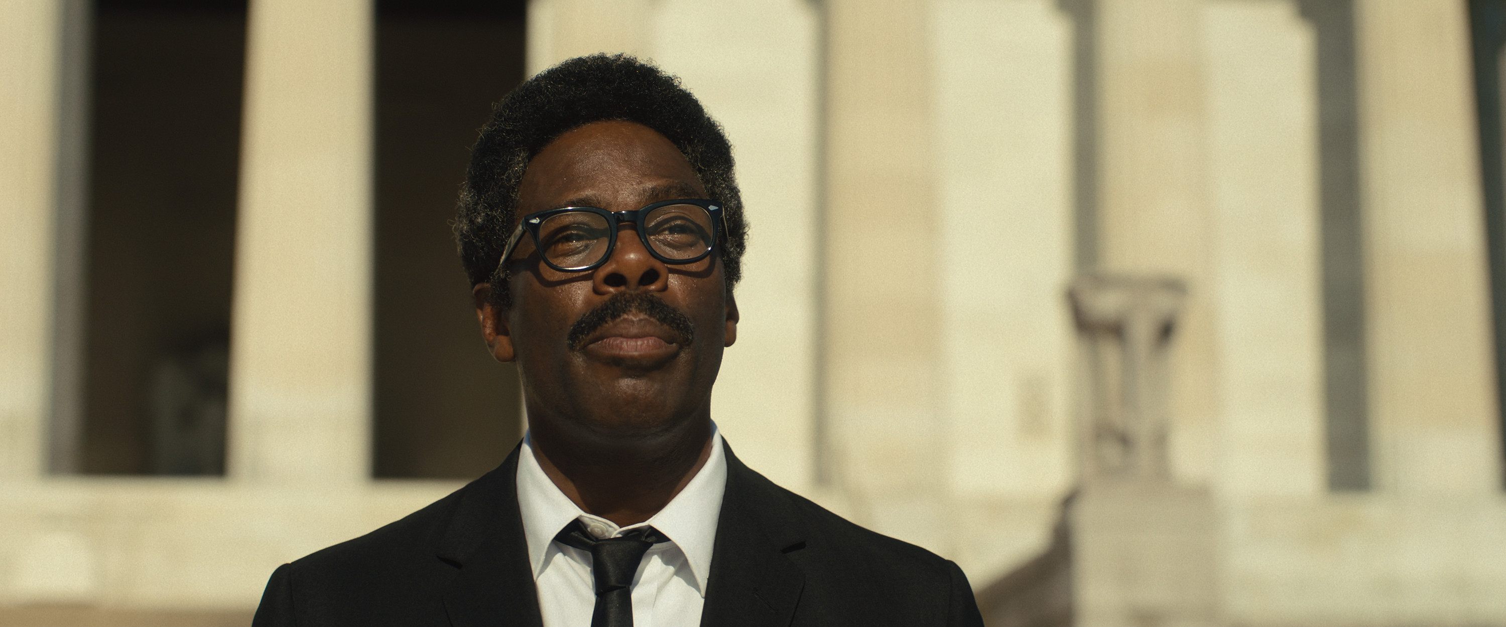 Colman Domingo as Bayard Rustin wearing a suit and glasses standing outside of a government building in the netflix movie rustin