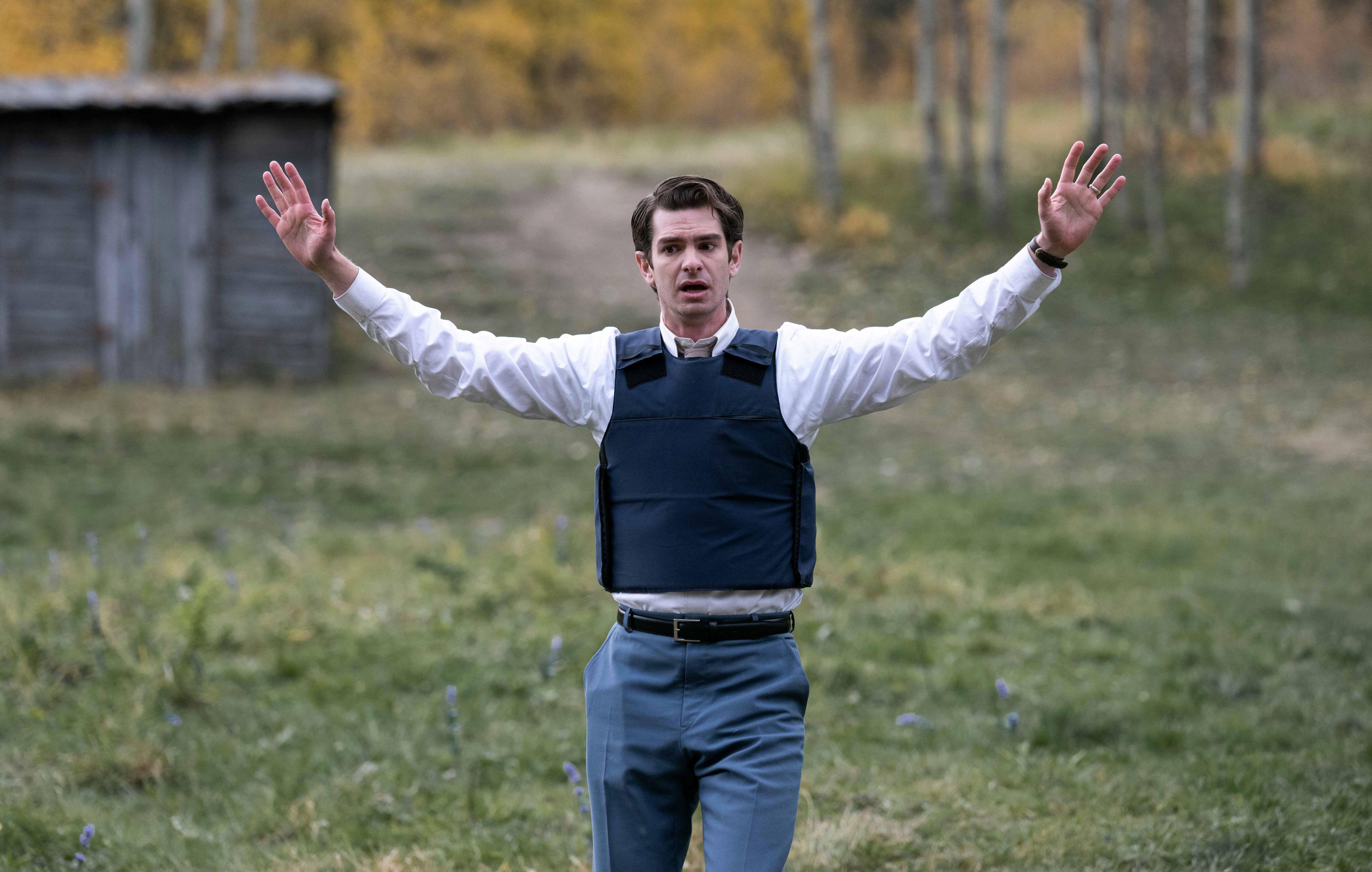 andrew garfield wears a bullet proof vest and waves his hands in the air in a rural field in under the banner of heaven