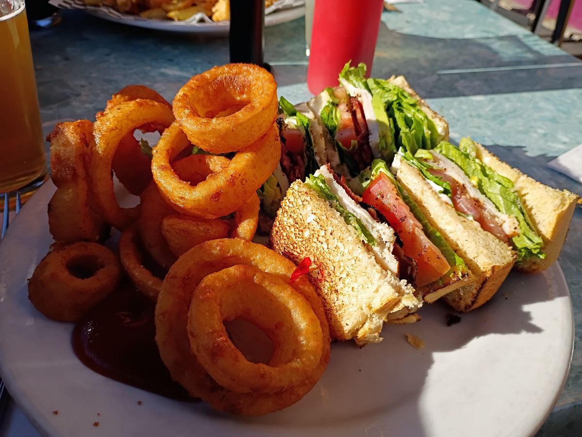 a white plate filled with large onion rings and a small sandwich in the shade outside on a sunny day