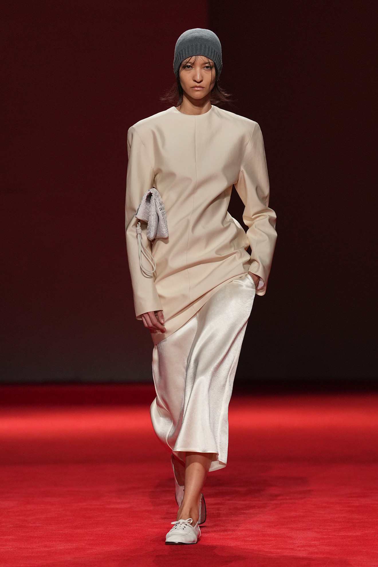 A Bottega Veneta model walking in the F/W 26 show wearing a gray beanie, a cream top, a cream skirt, and white flats.