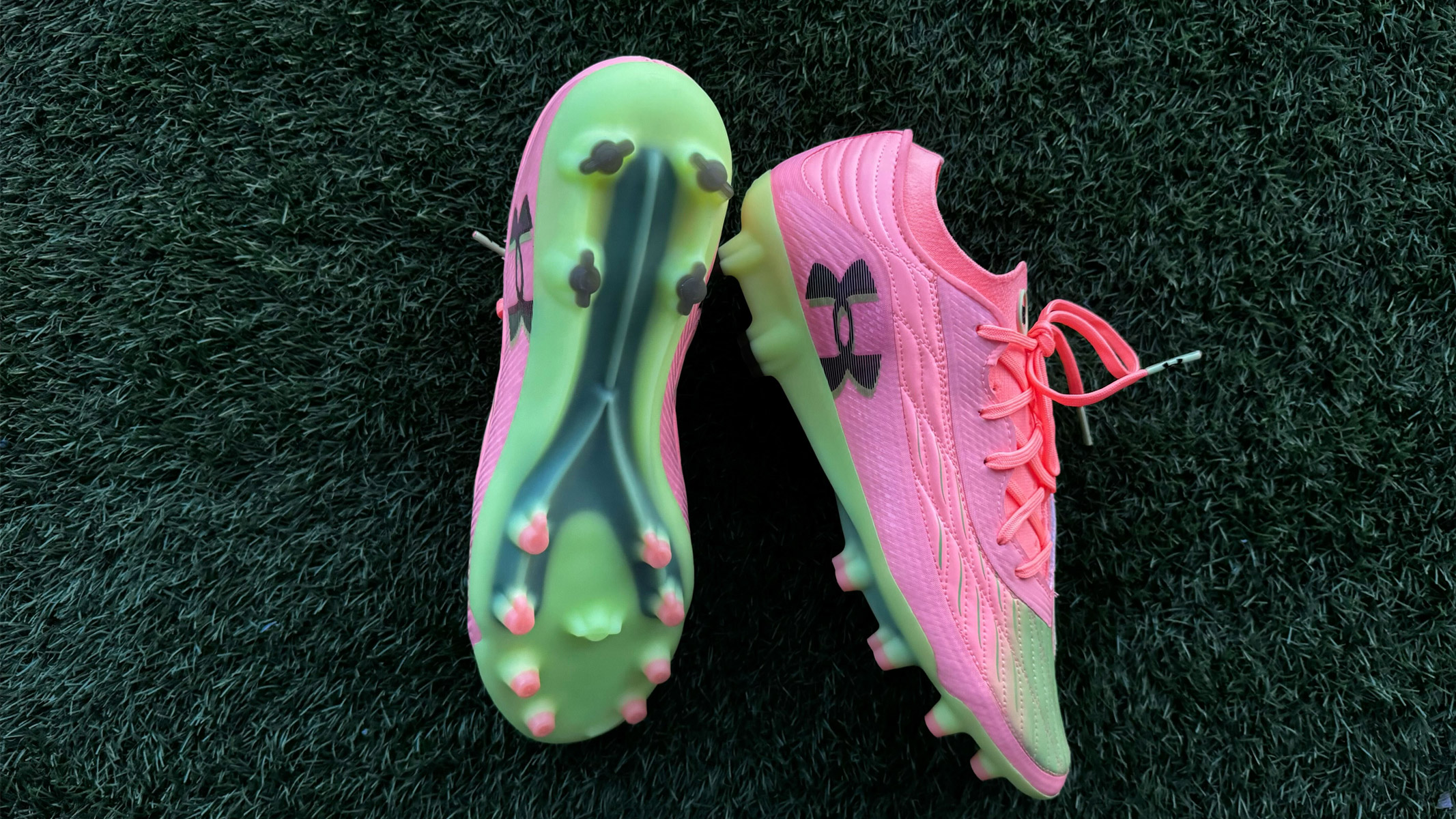 A look at the side and under-side of pink and green football boots with moulded studs, sitting on green artificial grass. Under Armour Clone Magnetico Elite 4.0