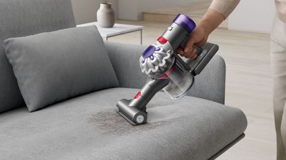 Hand holding the Dyson Car+Boat handheld vacuum, vacuuming gray sofa with small dirt patch in front of neutral living room.