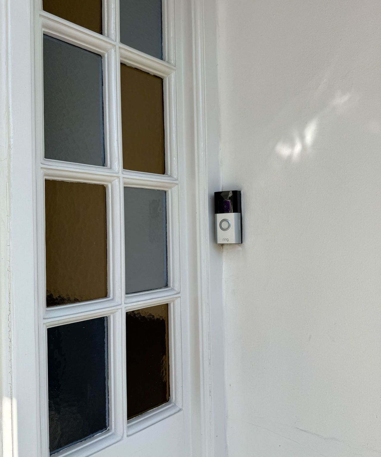 The Ring Battery Doorbell Plus mounted on a white painted wall, next to a yellow and blue paneled stain-glass window to the left of it.