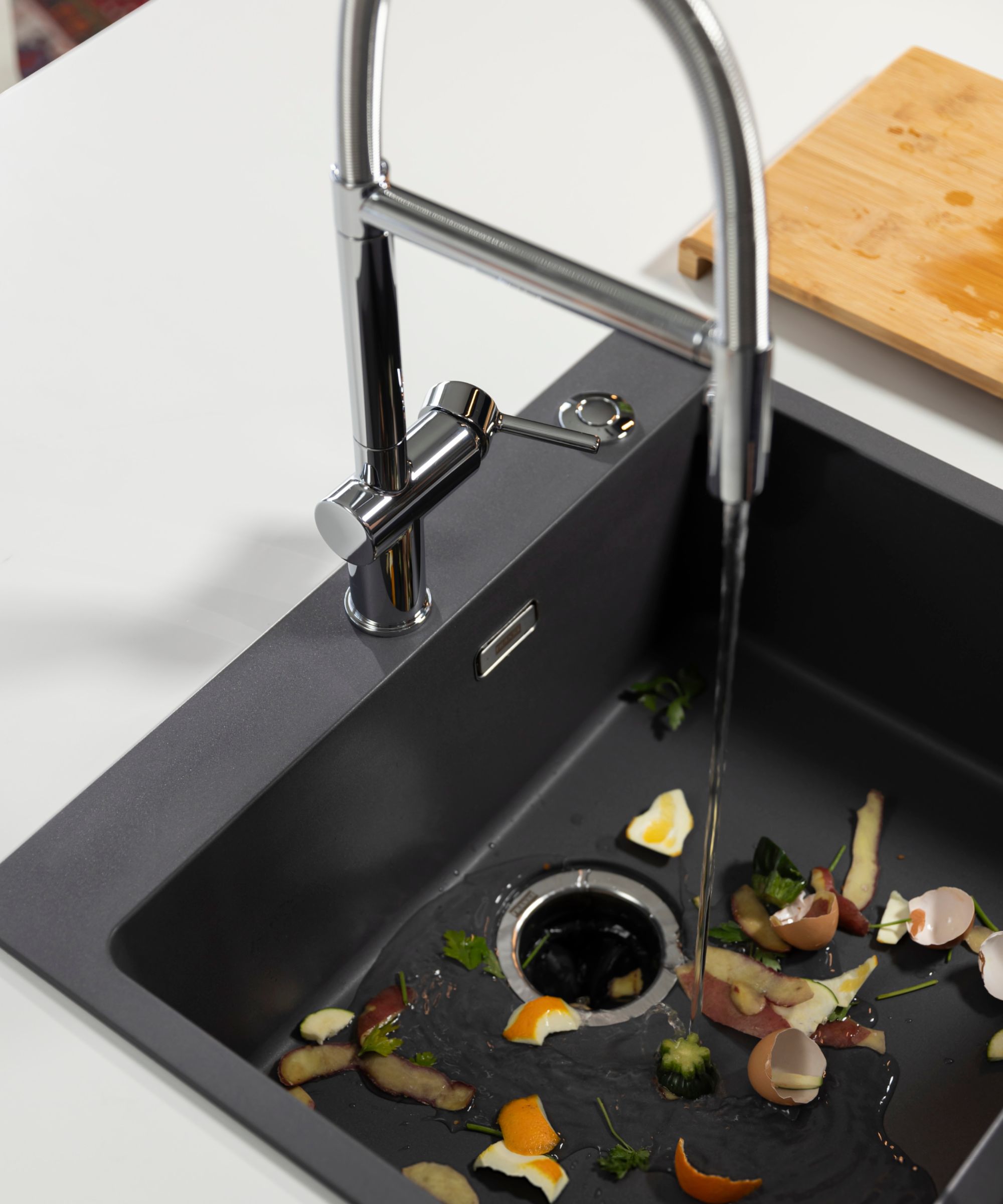 A modern kitchen sink with a chrome tap running water over food scraps including eggshells, vegetable peelings and citrus rinds, ready to be processed by a waste disposal unit.