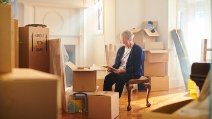 An older woman looks at a photo in her living room, surrounded by moving boxes.