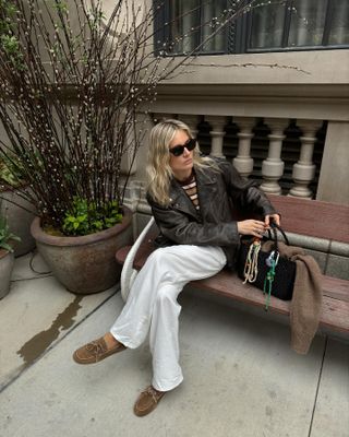 British fashion influencer Lucy Williams sits on a bench in NYC wearing a brown distressed leather jacket, Linda Farrow sunglasses, striped t-shirt, relaxed white pants, Little Liffner woven tote bag with charms, and tan suede loafers