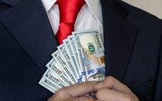A man in a navy blue suit and red tie tucking 100-dollar bills into his suit pocket