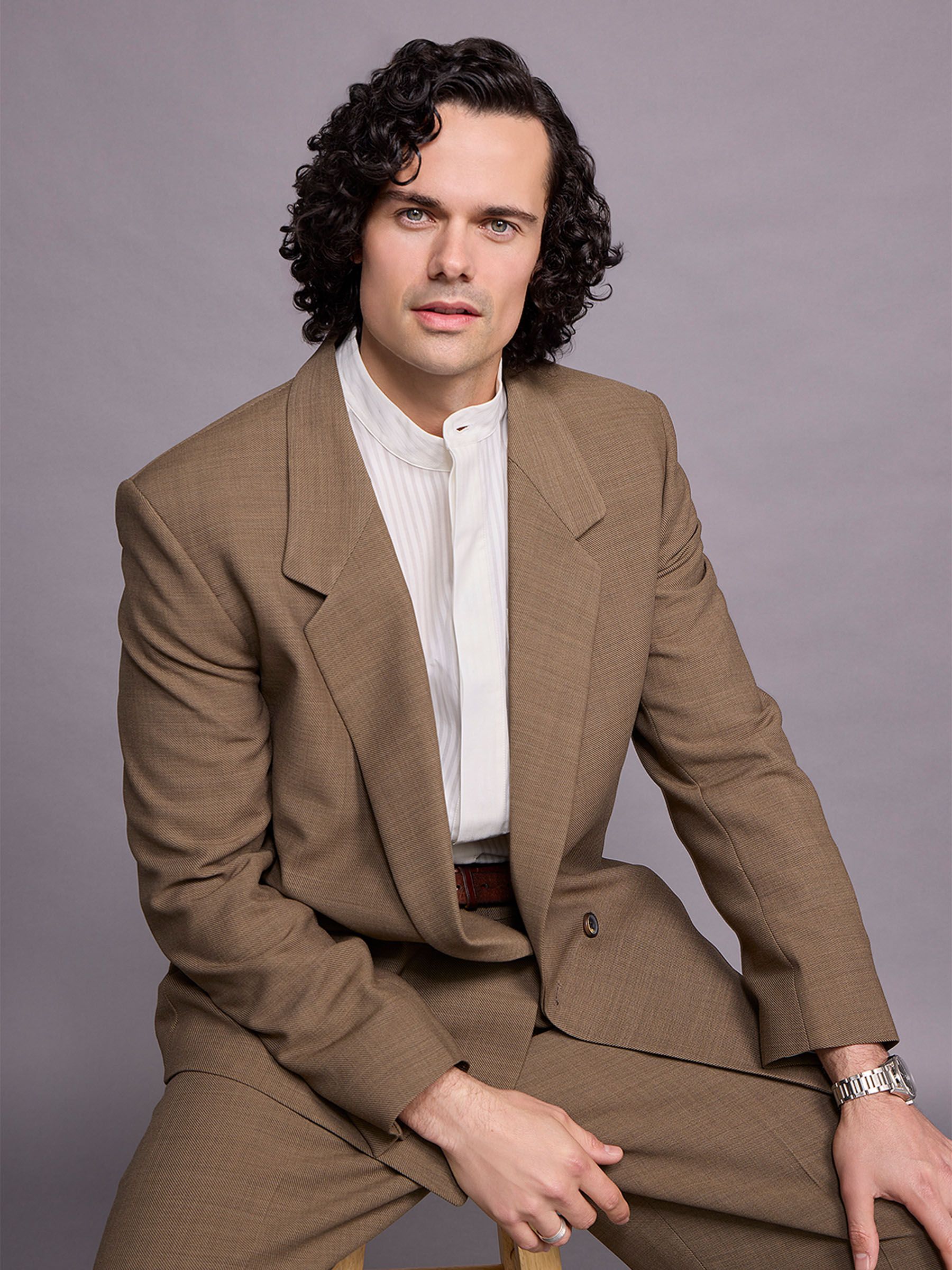 Actor Jamie Roy is sitting on a stool in front of a gray backdrop wearing a brown suit with a white tuxedo shirt.