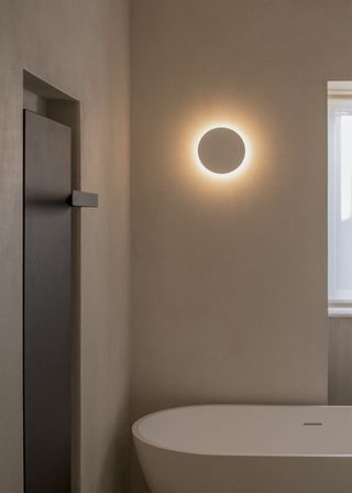 A small light fixture illuminated on a wall in a circular shape. Underneath it is a bath tub.