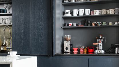 Dark grey kitchen cabinet with bi-fold doors open to show shelves full of mugs and a worktop coffee station set up