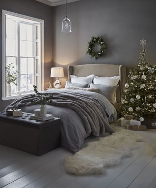 A dark grey bedroom with Christmas wreath on wall, Christmas tree, light wooden floorboards and grey velvet ottoman