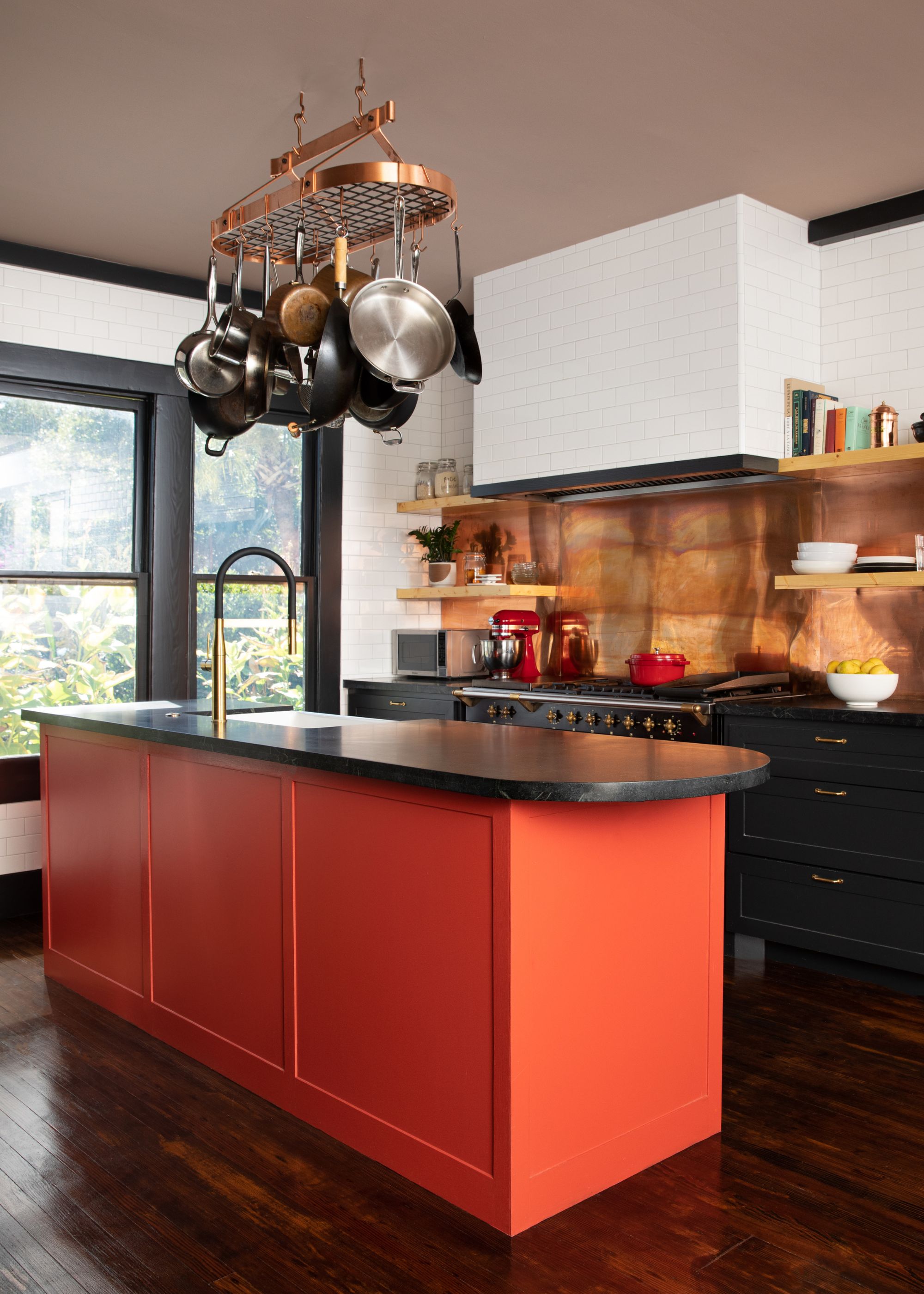 A small kitchen with a bright island and a copper splashback as well as pans including copper pans hanging above the island