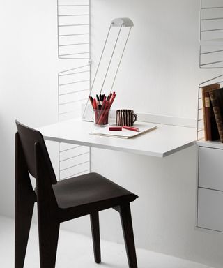 White String Furniture desk and shelving wall mounted unit, black desk chair, desk surface covered in pens and books.