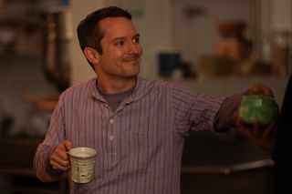 Elijah Wood as Walter wearing a striped shirt and smiling holding a coffee cup in YELLOWJACKETS