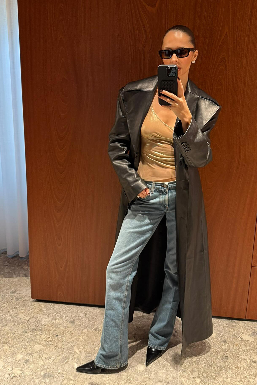 A woman wearing a black leather coat, tan tank top, light blue jeans and black pointe shoes