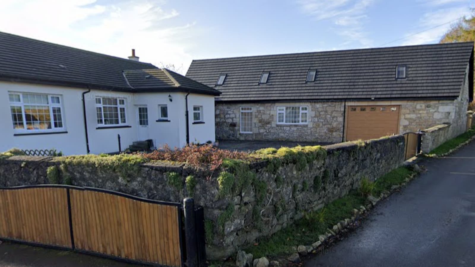 A wooden gate and stone fence and a single storey stone house next to a white brick house