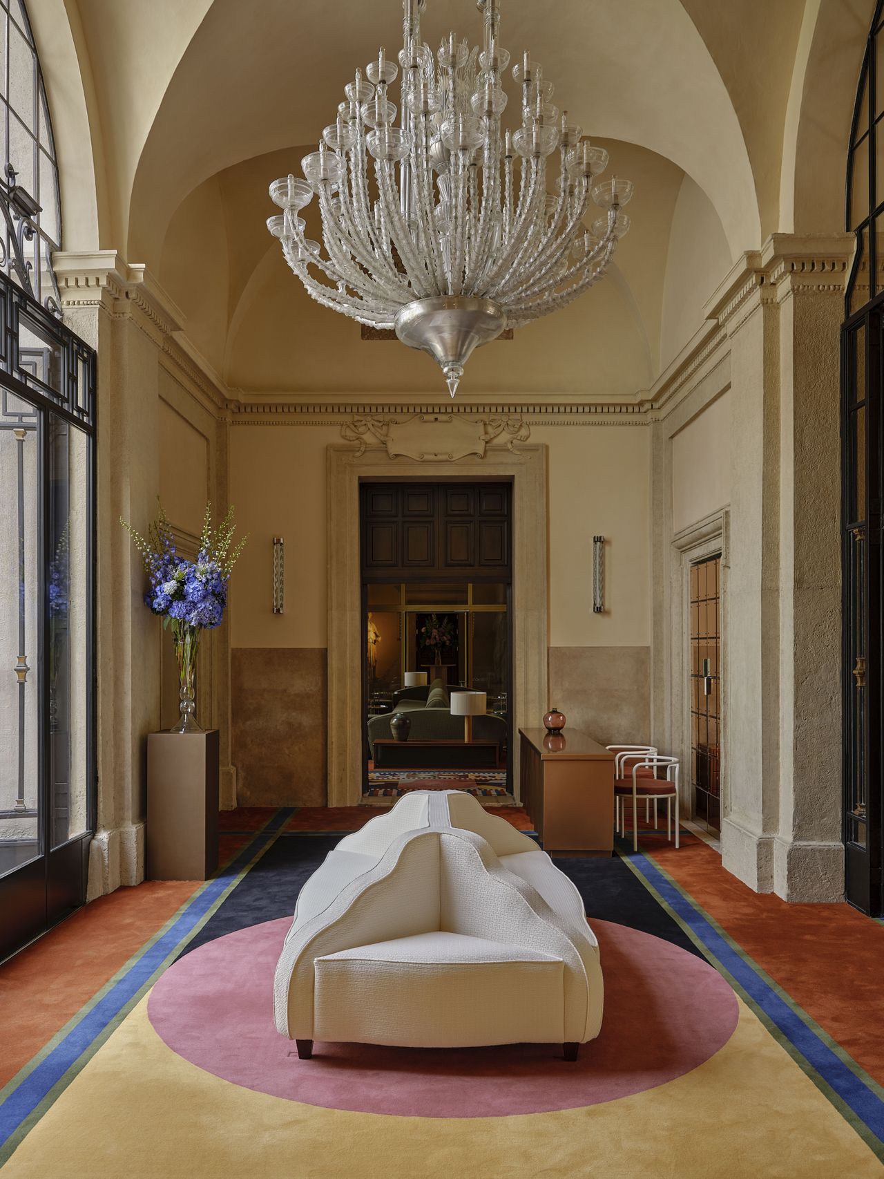 A whimsically reinvented palace hallway features a colorful rug made of contrasting shades and shapes, a Murano glass chandelier, flowers, and other vintage-inspired collectibles.