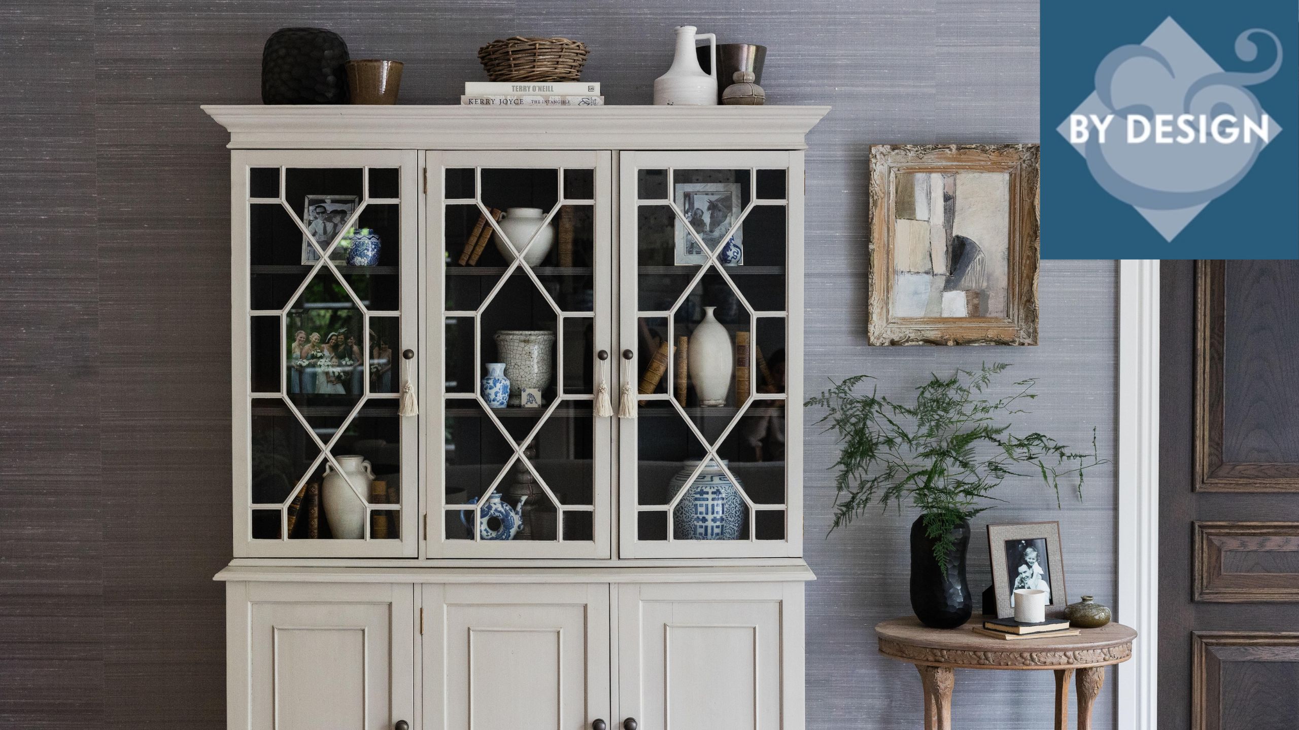 Antique large white dresser filled with ornaments, textured gray wallpaper behind, side table decorated with plant and pictures to the right, artwork hung above