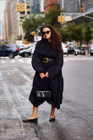 An elegantly dressed woman wearing a black belted dress and black flats stands on a rainy street in new York city.