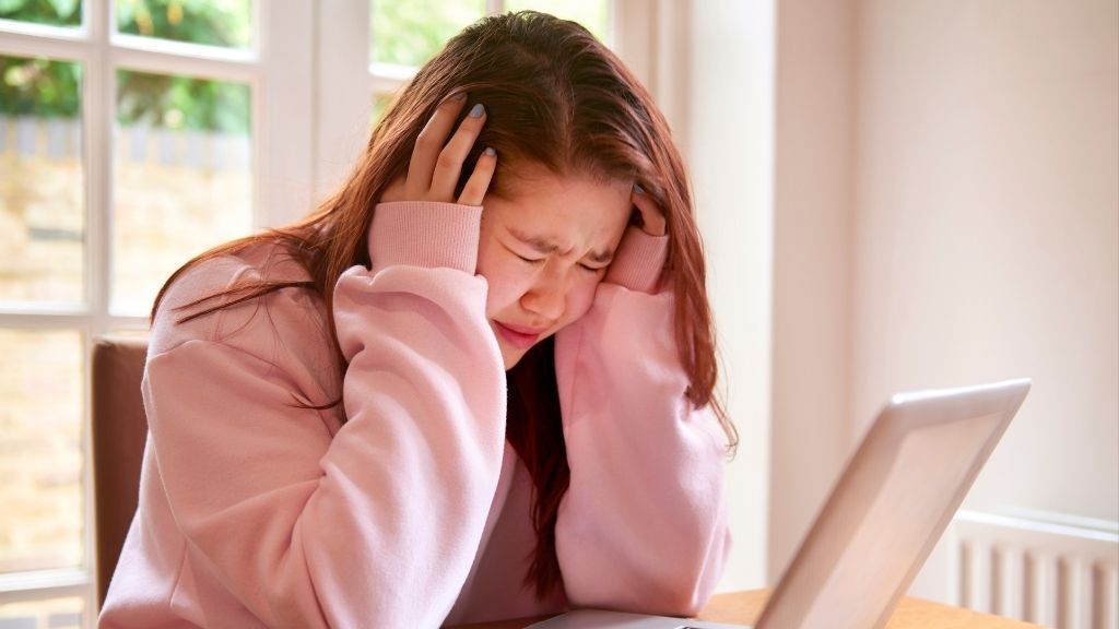 A young woman wearing a pink sweater holds her head in both hands and scrunches up her eyes in pain, as if experiencing a headache