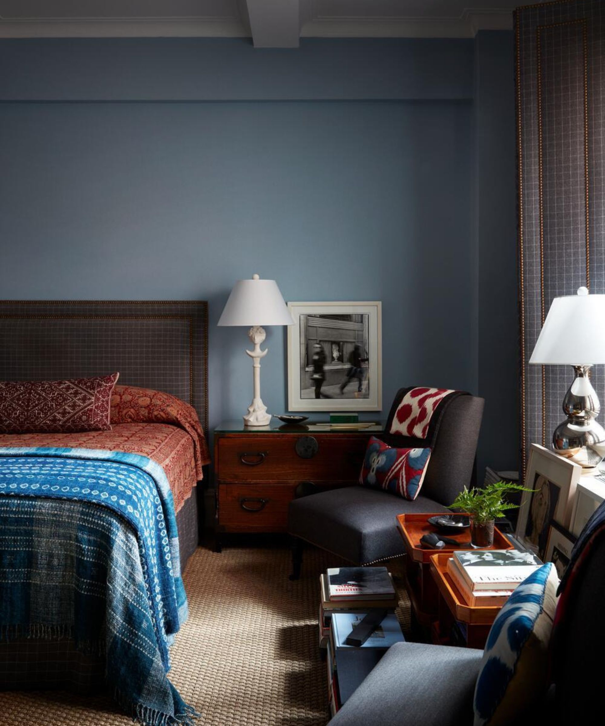 A moody bedroom with dusty blue walls, featuring a bed with layered blue and red textiles, a vintage wooden chest as a nightstand, and a mix of framed art and patterned pillows