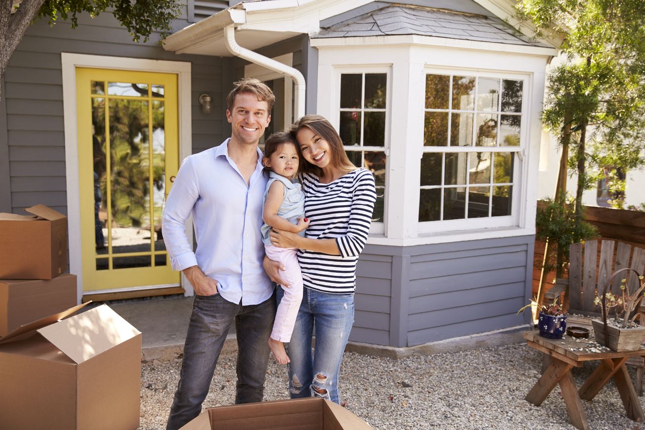 A young couple stands in front of their newly-acquired house, holding their young daughter, with moving boxes to the side.