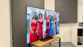 65-inch LG C4 TV photographed at a slight angle on a wooden stand. On the screen is an image of a group of women in brightly coloured dresses.